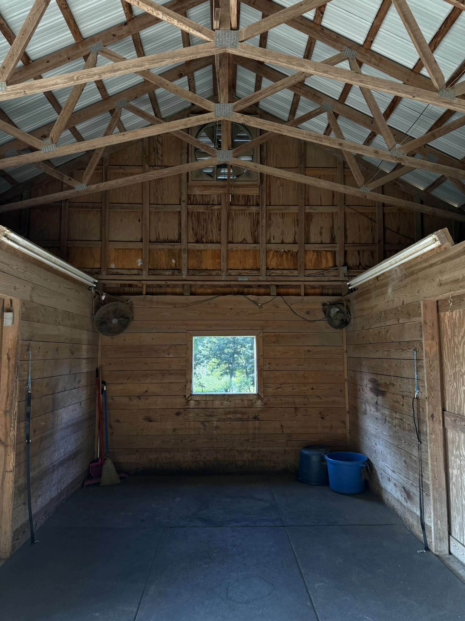 Empty wooden barn interior with a small square window showing green trees outside, two fans mounted on walls, cleaning tools in left corner, and two blue buckets on the right.