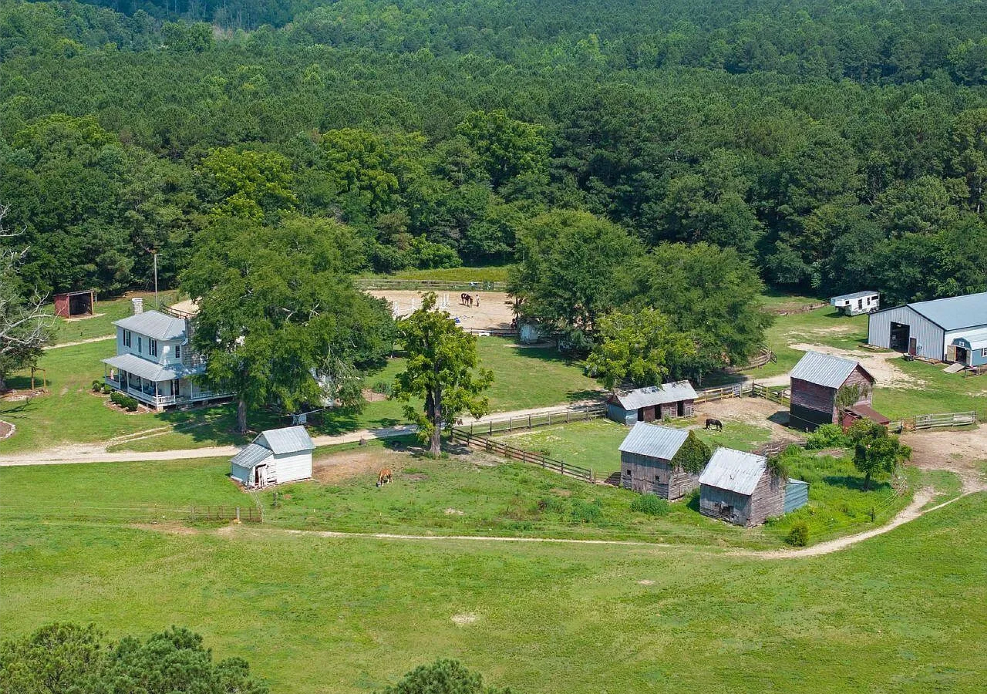 Aerial view of a farm with several barns, fenced pastures, a large white house with a porch, horses, and dense forest in the background.