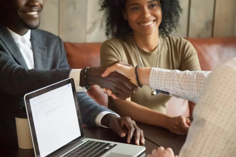 Two people shaking hands across a table with a laptop, while a smiling woman watches.