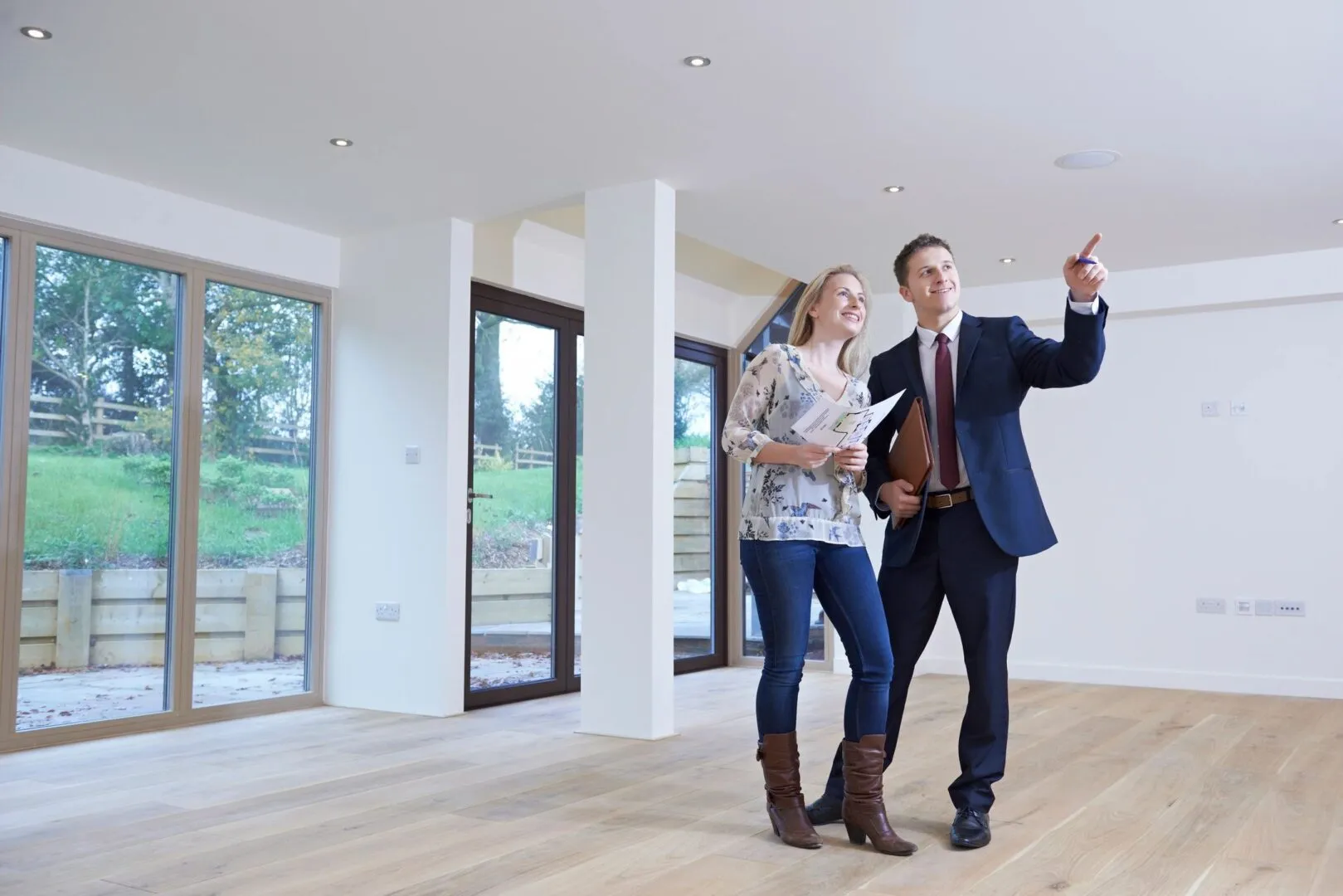 Real estate agent showing a woman an empty bright room with large windows and wooden floor.