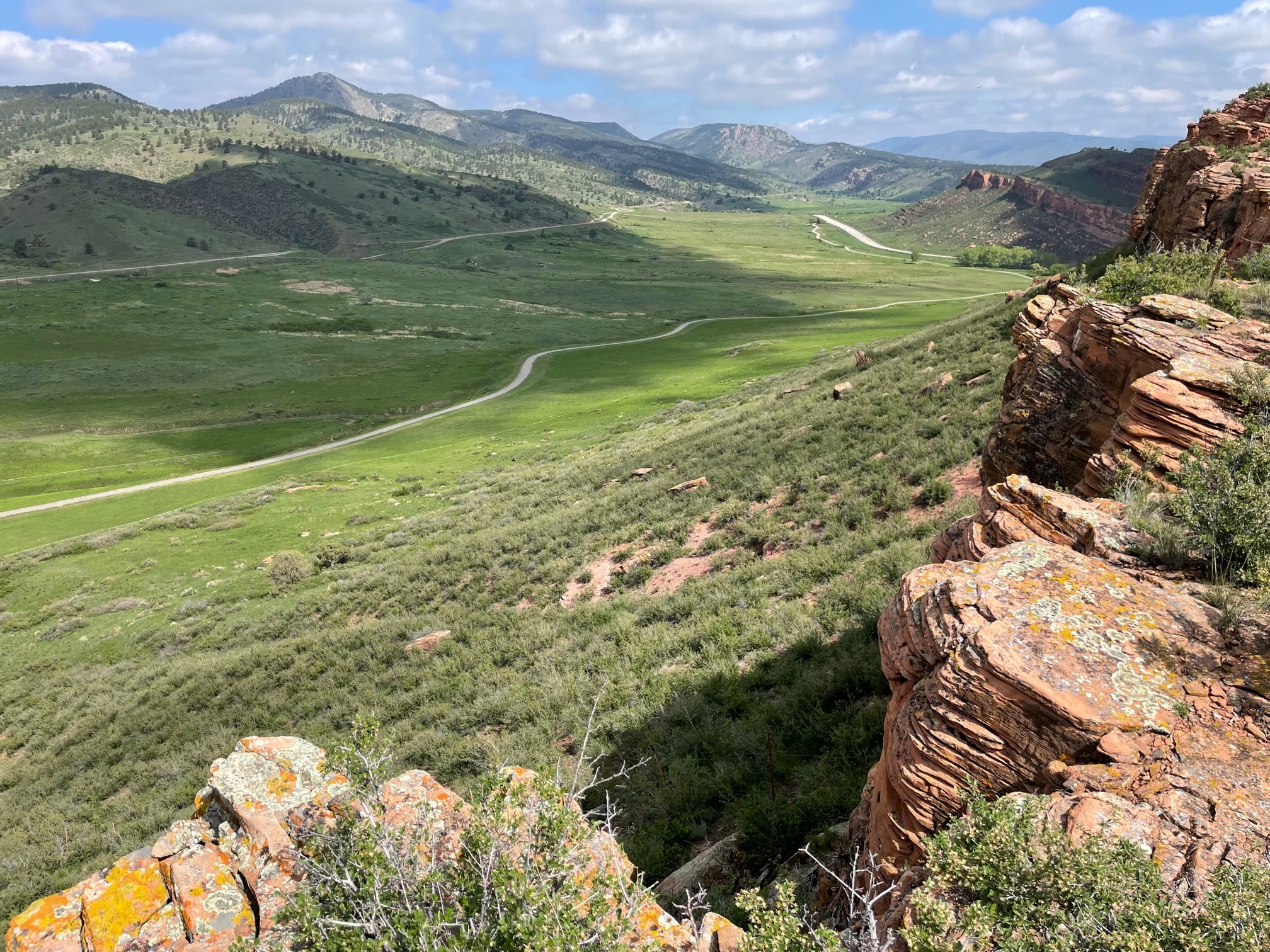 Green valley with winding road, rocky cliffs on right, and mountains under a blue sky with clouds.