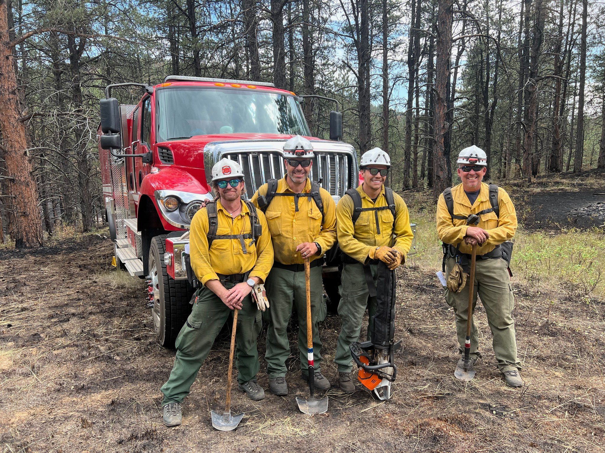 Four firefighters in yellow shirts and helmets stand with tools in front of a red fire truck in a forest.