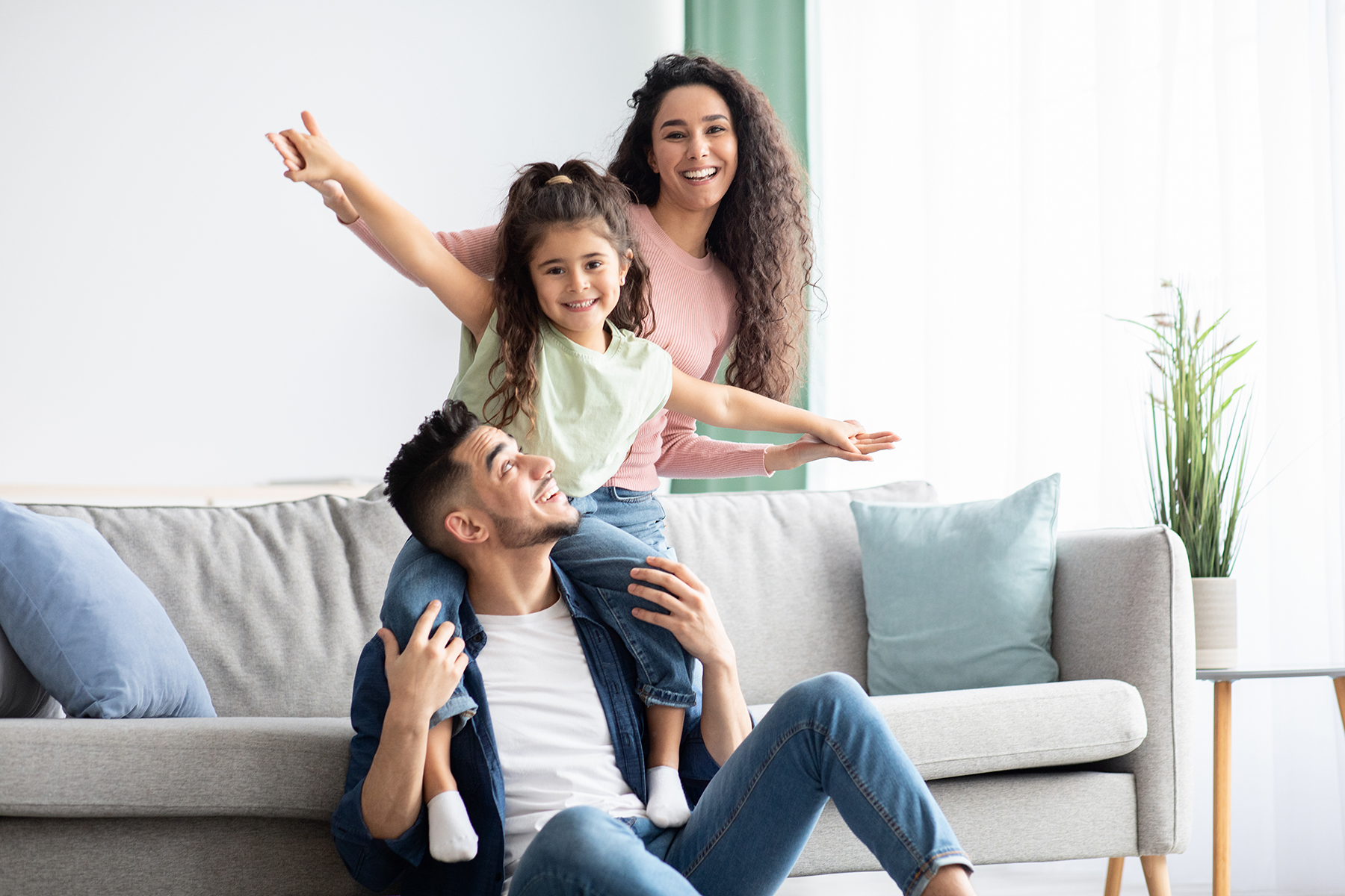 Smiling family with father holding daughter on shoulders and mother behind on living room sofa.