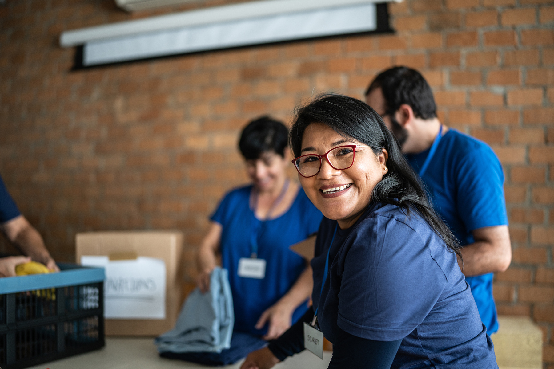 Smiling woman with red glasses volunteers packing donations at a table with two other volunteers.