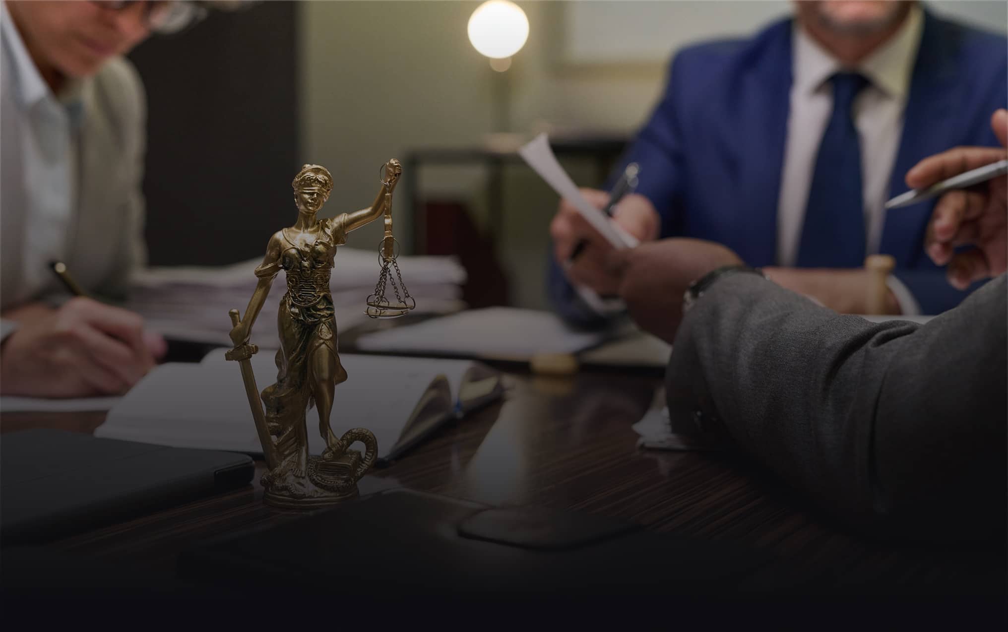 Bronze Lady Justice statue on a desk with people in business attire reviewing documents in the background.
