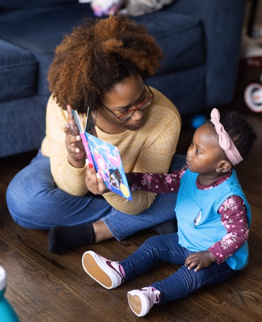 At Small Magic, an adult reads with a young child seated on the floor, while the child wears a blue LENA device vest.