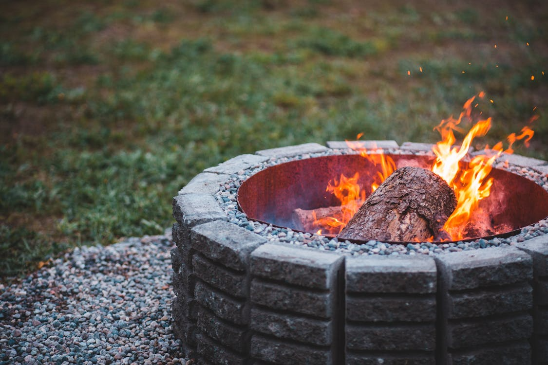 Stone fire pit with burning logs and flames on a gravel base in a grassy outdoor area.