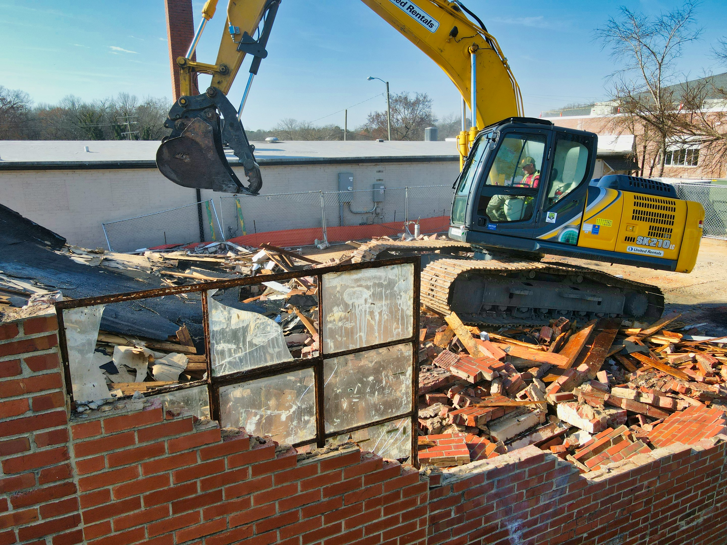 Yellow excavator demolishing a brick building with rubble and broken window panes in the foreground.