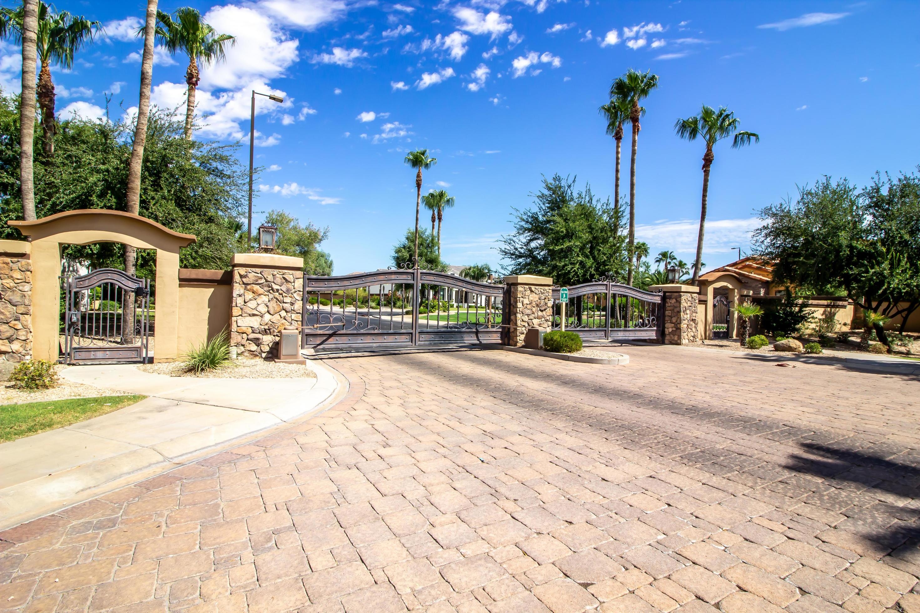 Stone gate entrance with wrought iron gates and palm trees under a partly cloudy blue sky.