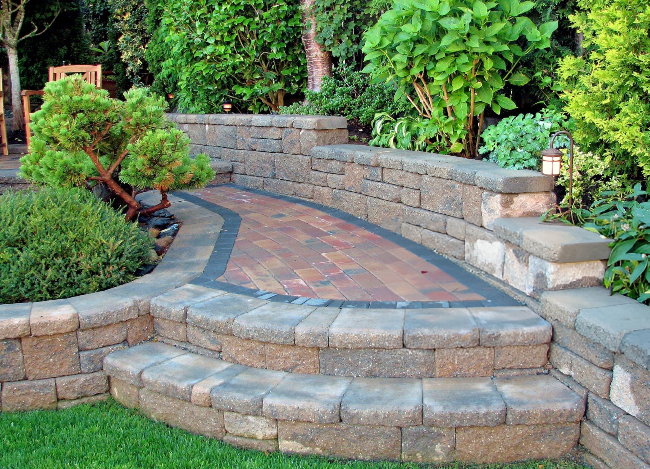 Curved brick pathway with low stone walls and steps surrounded by green shrubs and plants.