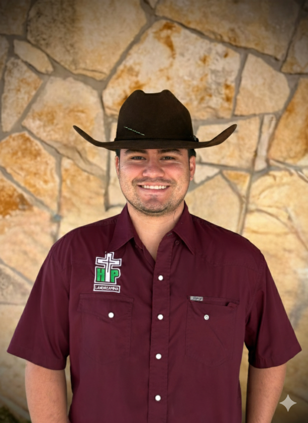 Smiling man wearing a brown cowboy hat and maroon shirt with landscaping logo standing in front of a stone wall.