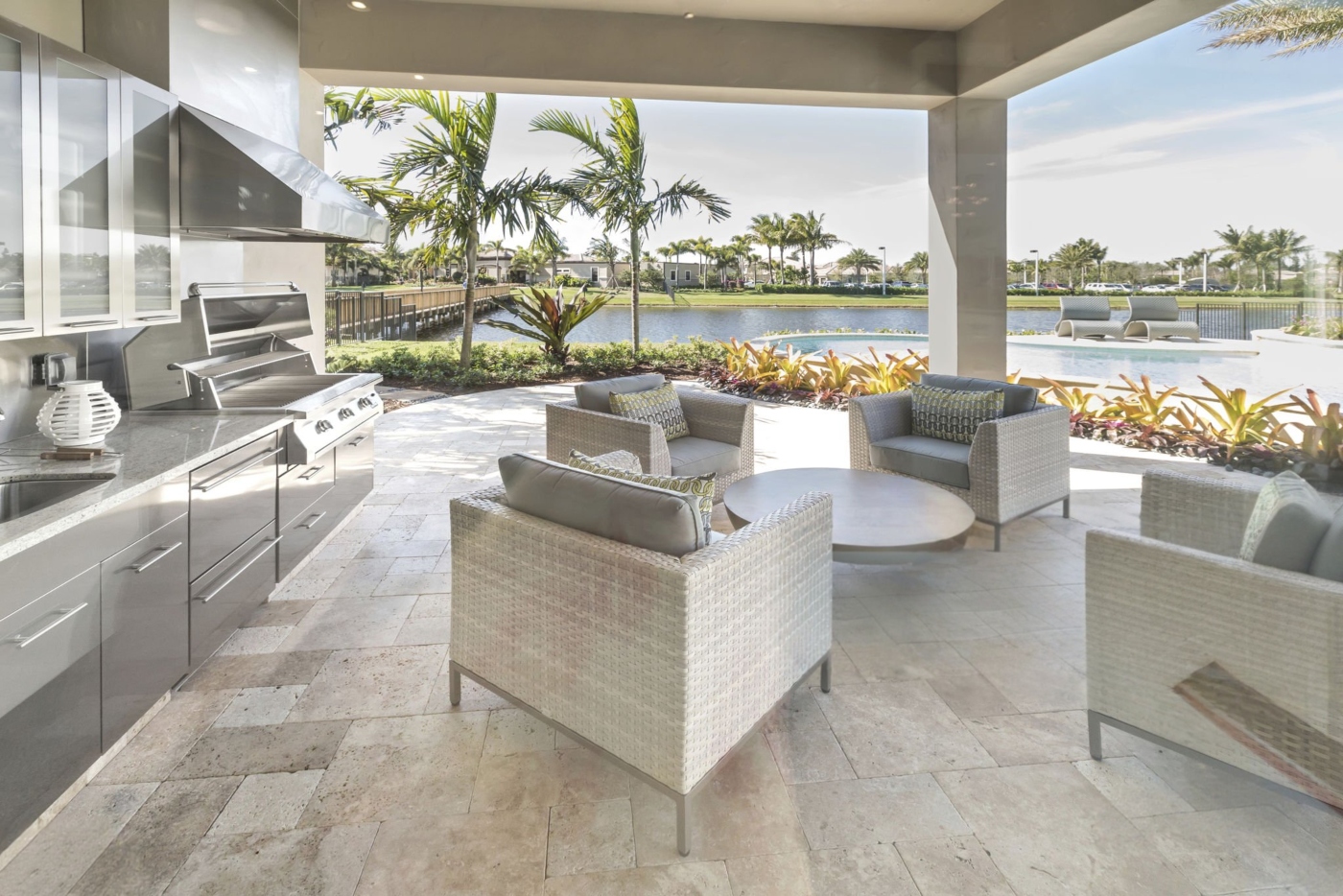 Modern outdoor kitchen and seating area beside a pool with palm trees and water view.