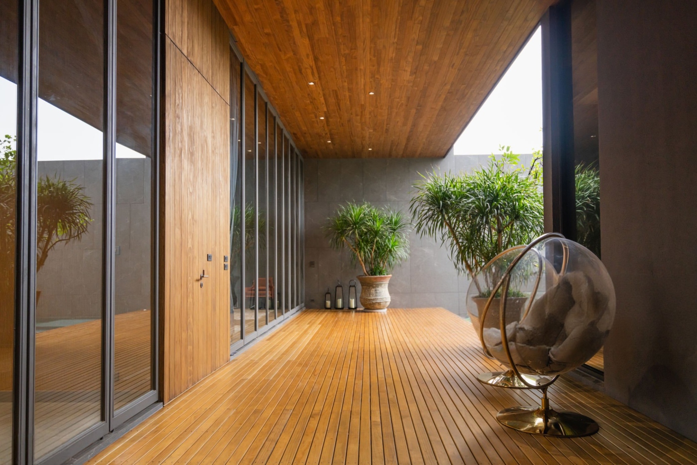 Wooden deck area with glass wall, large potted plants, and a transparent hanging chair with cushions.