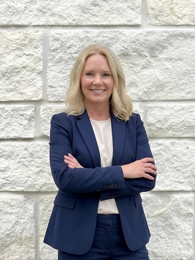 Smiling woman with blonde hair in a navy blue suit standing with arms crossed against a white stone wall.