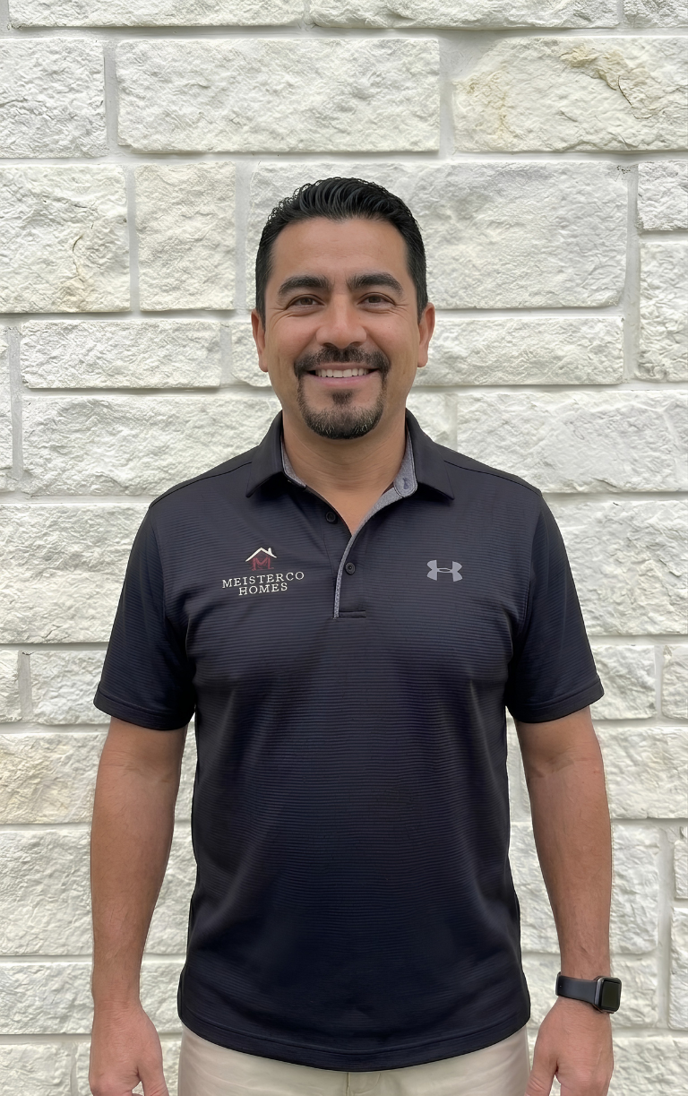 Smiling man wearing a black Meisterco Homes polo shirt and a smartwatch, standing against a stone wall.