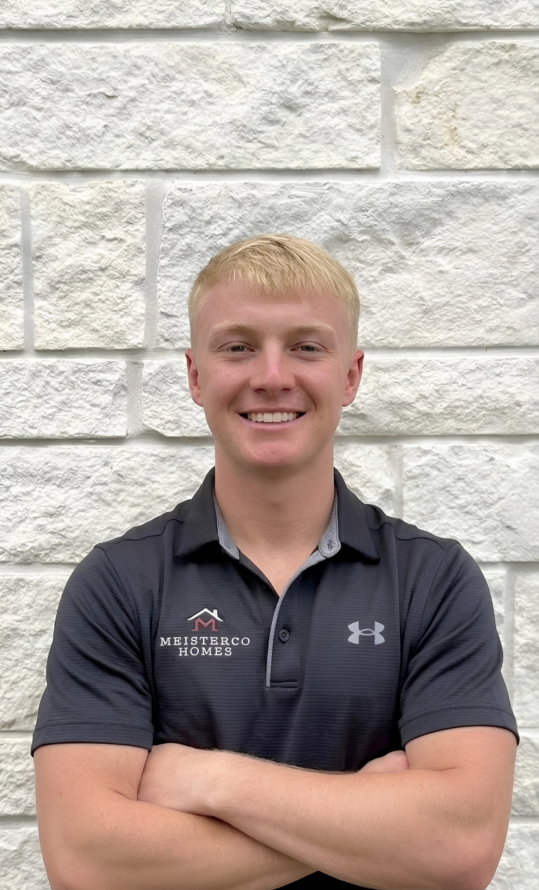 Smiling young man with blonde hair in a black Meisterco Homes polo shirt stands with arms crossed.