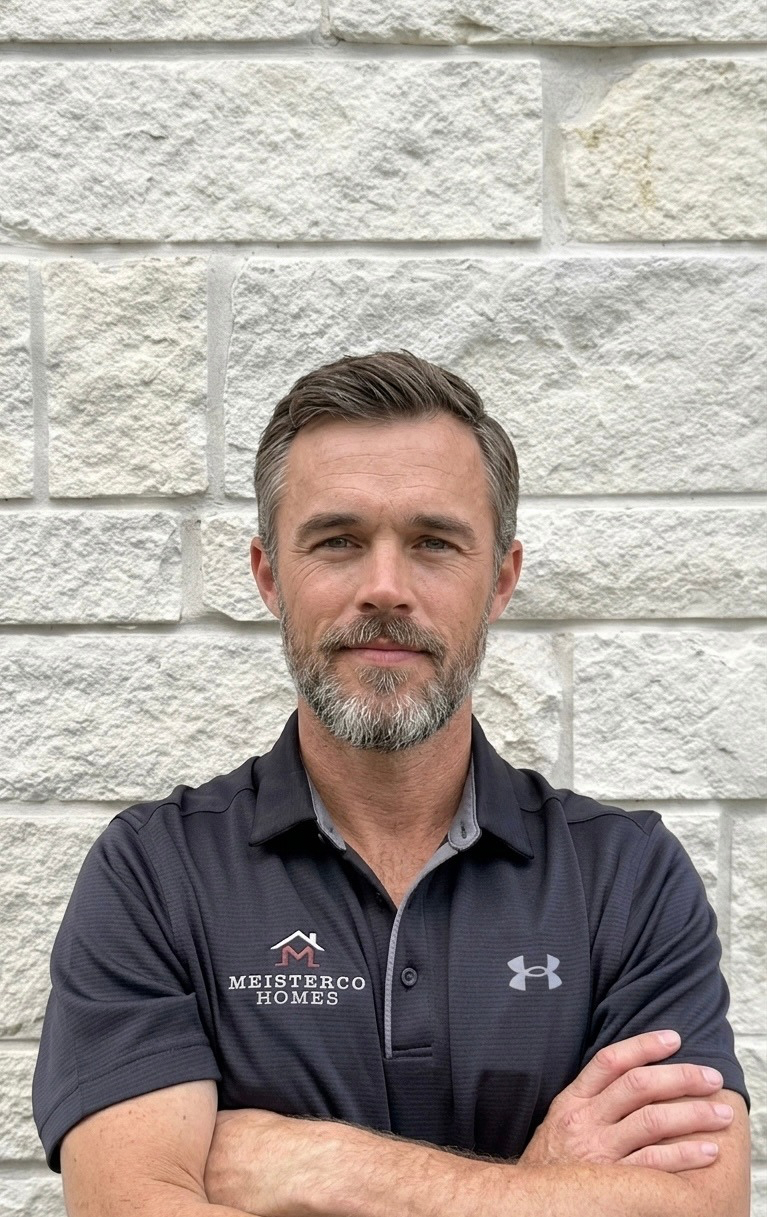 Man with short hair and beard, arms crossed, wearing a Meisterco Homes polo, standing against stone wall.