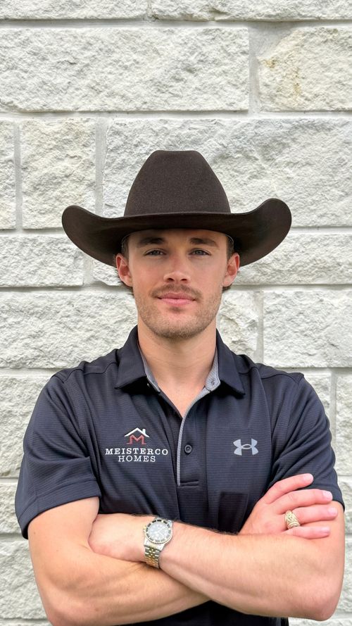 Man with mustache wearing black cowboy hat and Meisterco Homes shirt, arms crossed, standing by stone wall.