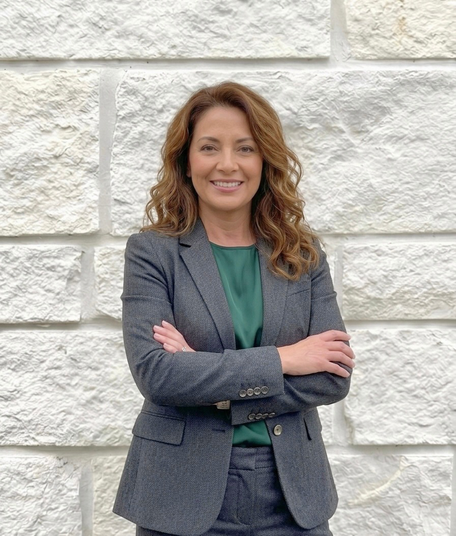 Smiling woman with wavy hair wearing gray suit and green blouse stands with arms crossed in front of white stone wall.