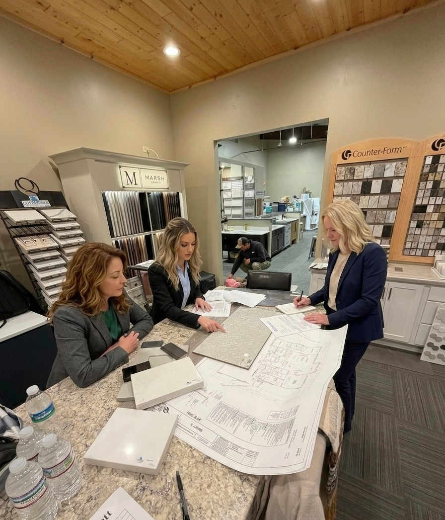 Three women in an office discussing floor plans and material samples on a table.