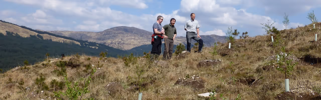 Three people standing on a grassy hill with mountains and a blue sky with clouds in the background.