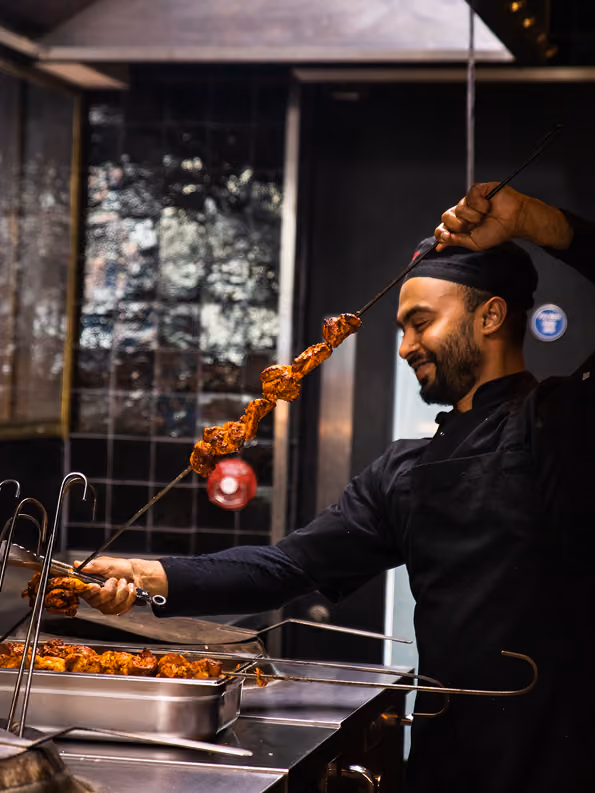 Chef in black uniform grilling seasoned chicken skewers on an open flame in a restaurant kitchen