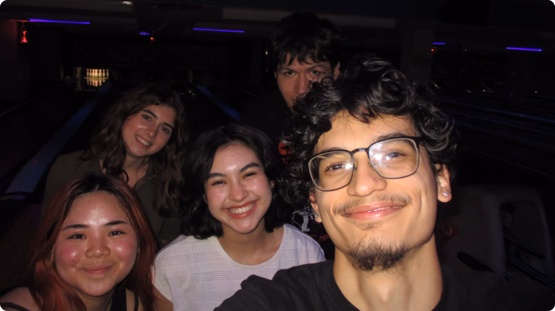 Group of five young people smiling and taking a selfie in a dimly lit bowling alley.