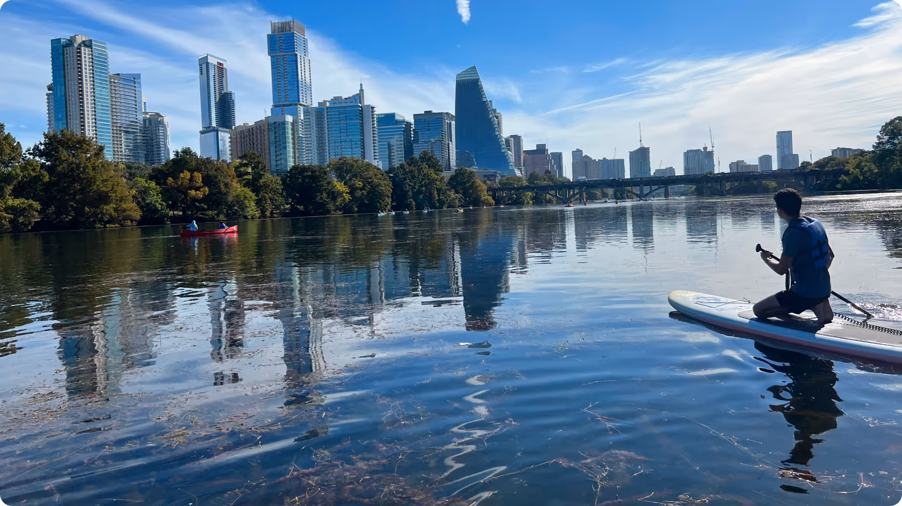 Person kneeling on a paddleboard paddling on a calm river with city skyscrapers and a bridge in the background under a blue sky.