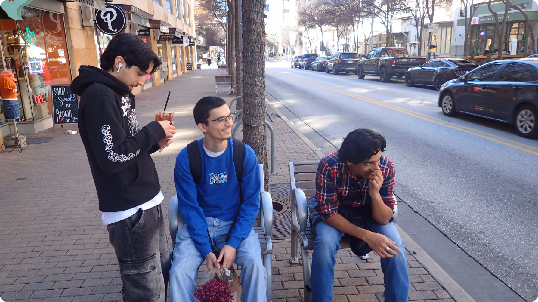 Three young men on a city sidewalk, one standing with a drink and two sitting on chairs near a tree-lined street with parked cars.