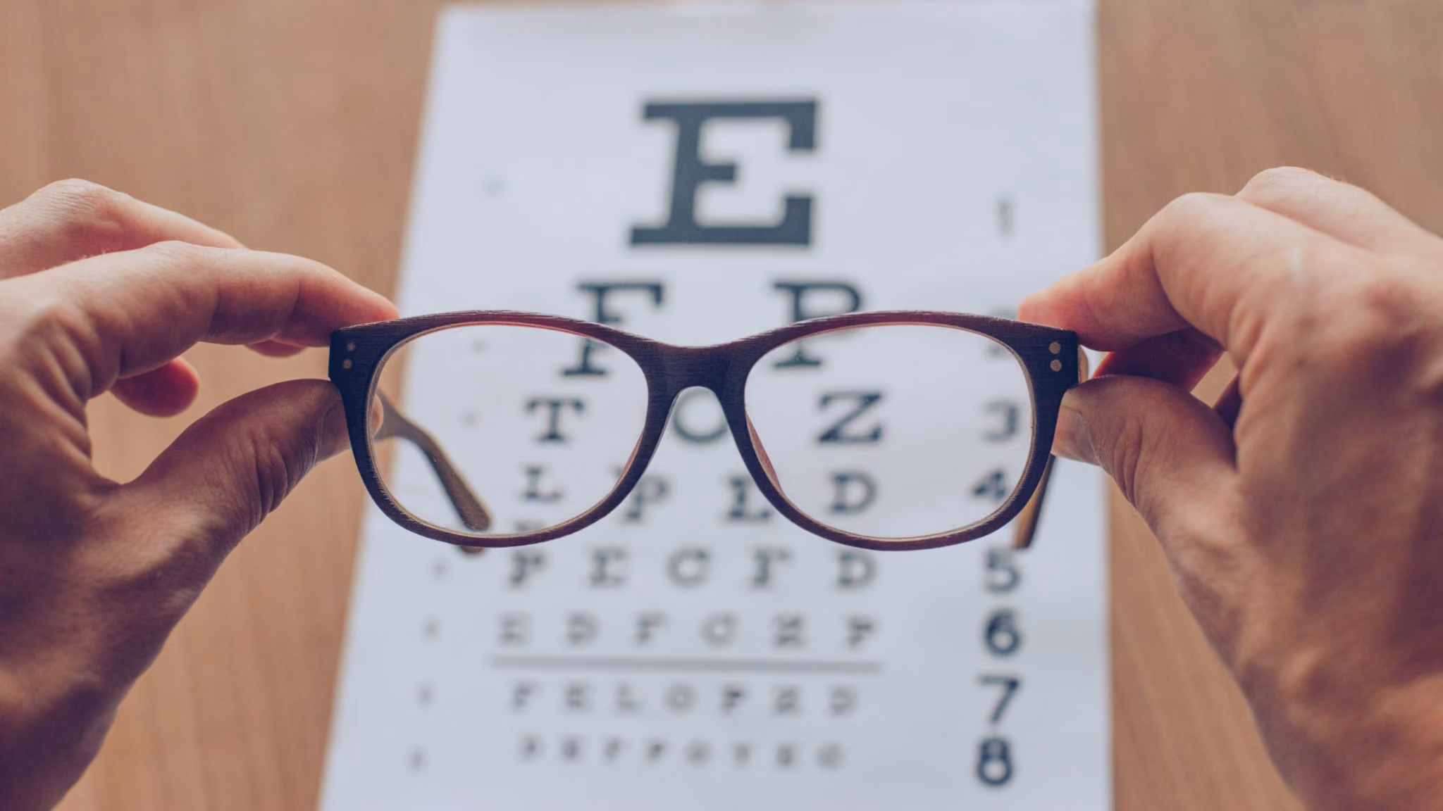 Hands holding eyeglasses focusing on a Snellen eye chart in the background.