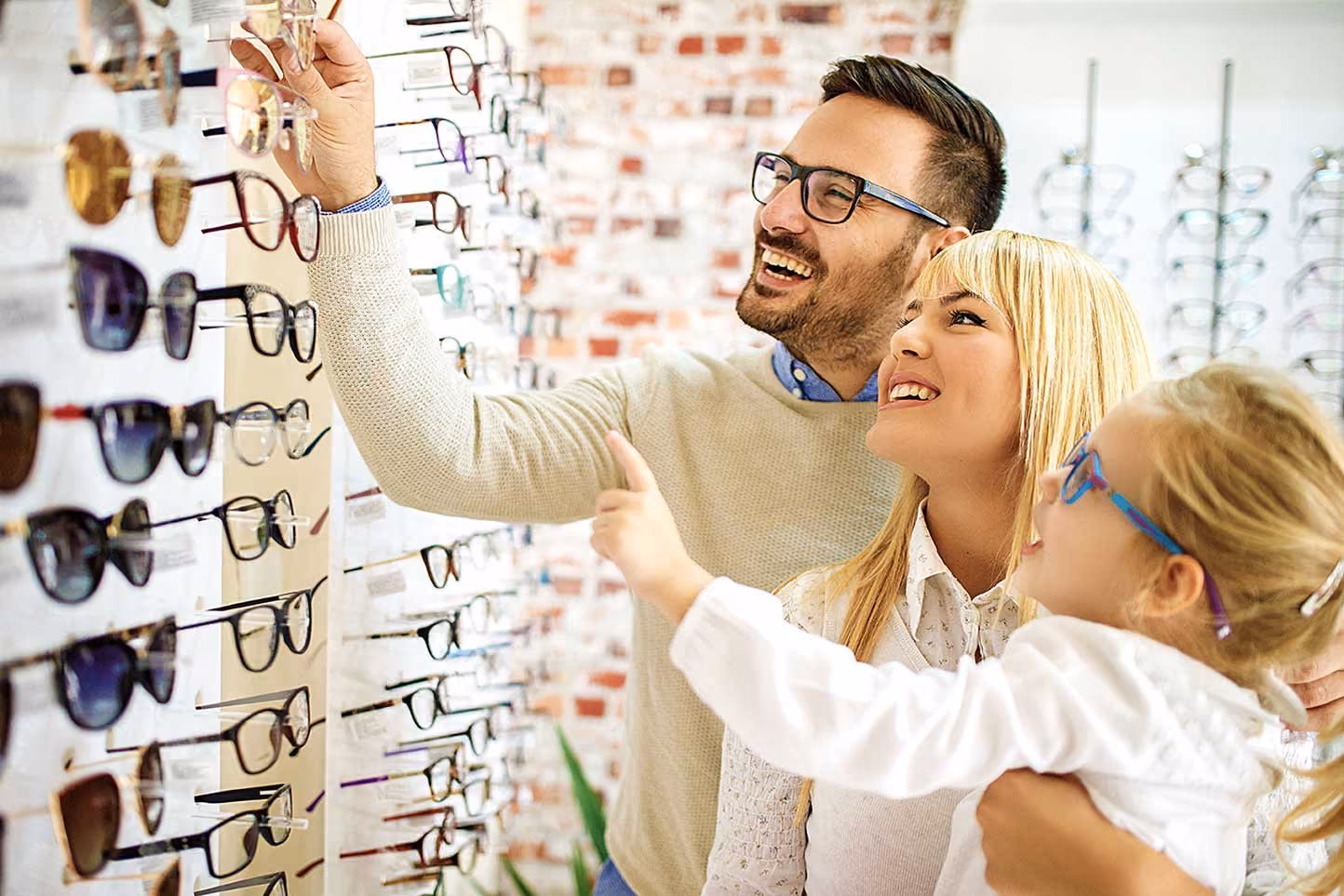 Smiling family of man, woman, and child trying on eyeglasses at an optical store display.