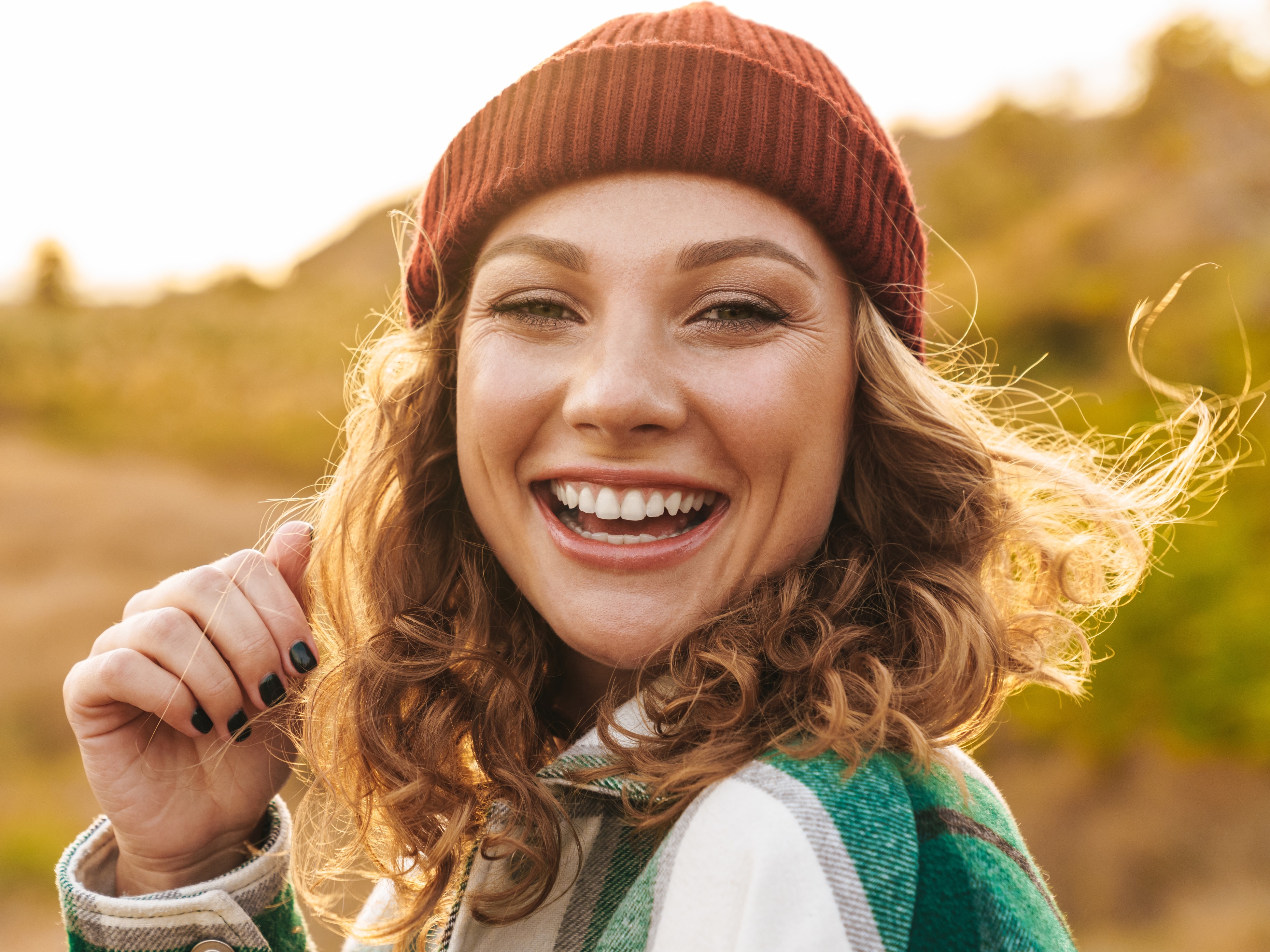 Smiling young woman with curly hair wearing a red knit beanie and green plaid jacket outdoors at sunset.