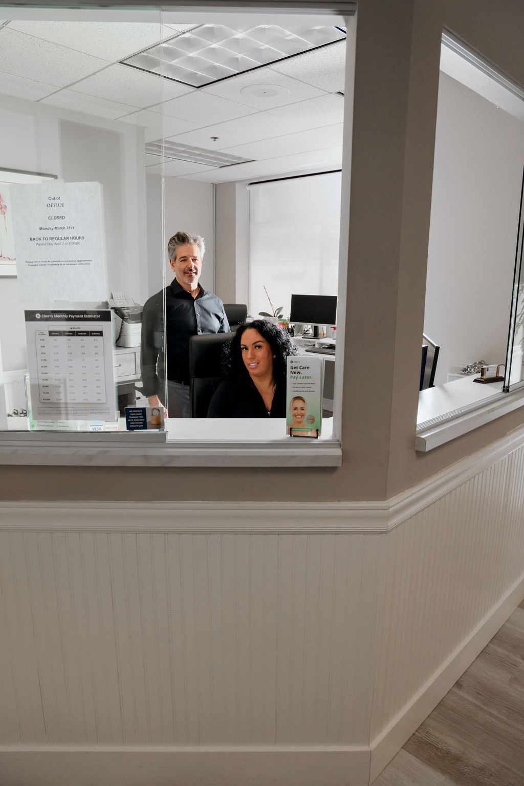 Two office workers behind a glass reception desk with signs and a computer in the background.