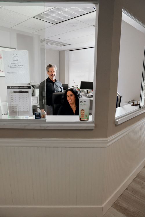 Two office workers behind a glass reception desk with signs and a computer in the background.