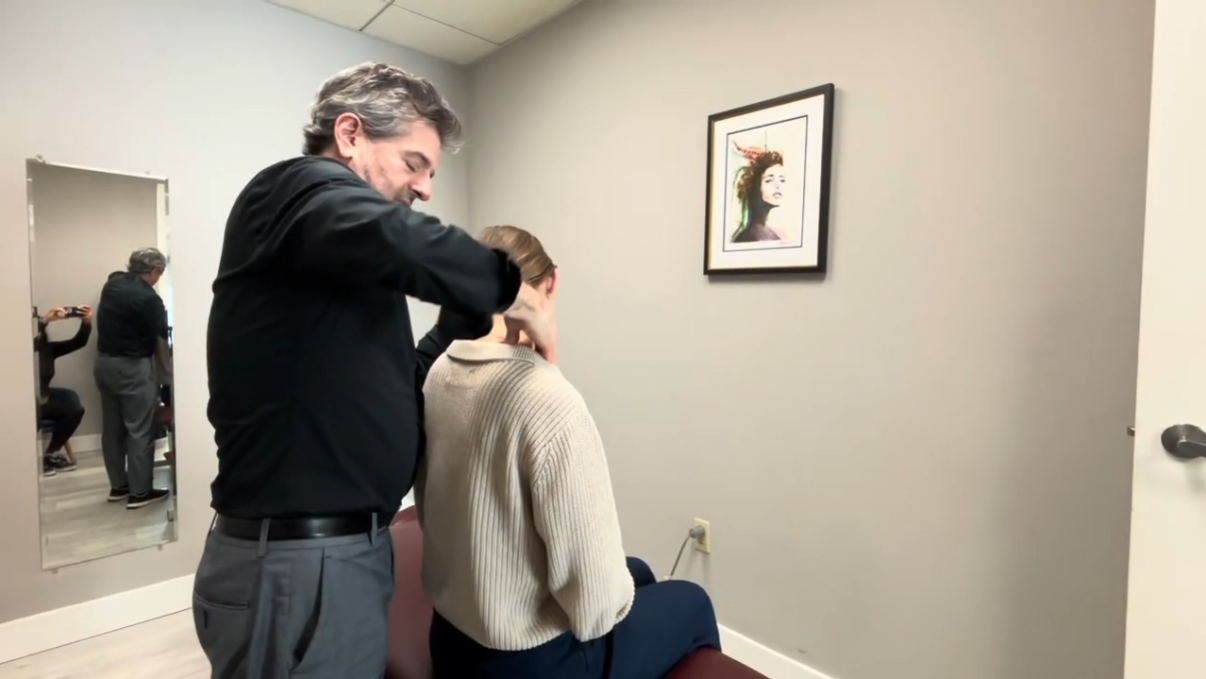 Man in black shirt giving a neck massage to a seated woman with a beige sweater in a minimally decorated room with a framed portrait on the wall.