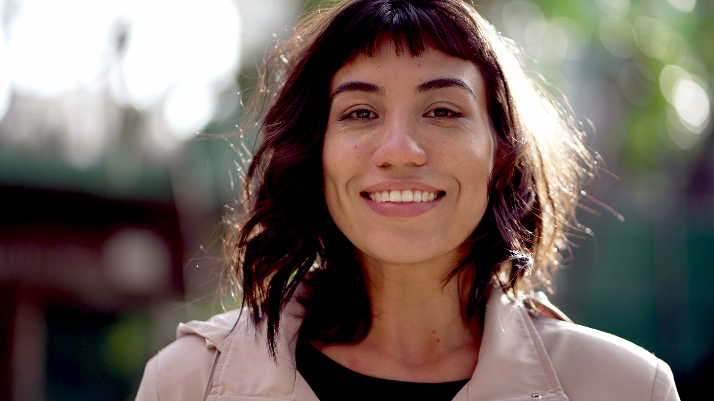 Smiling woman with short wavy hair and bangs wearing a beige jacket outdoors in soft daylight.