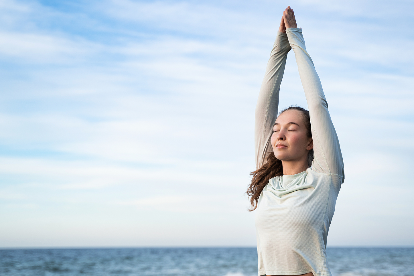Woman practicing yoga with arms raised and eyes closed by the ocean under a blue sky.
