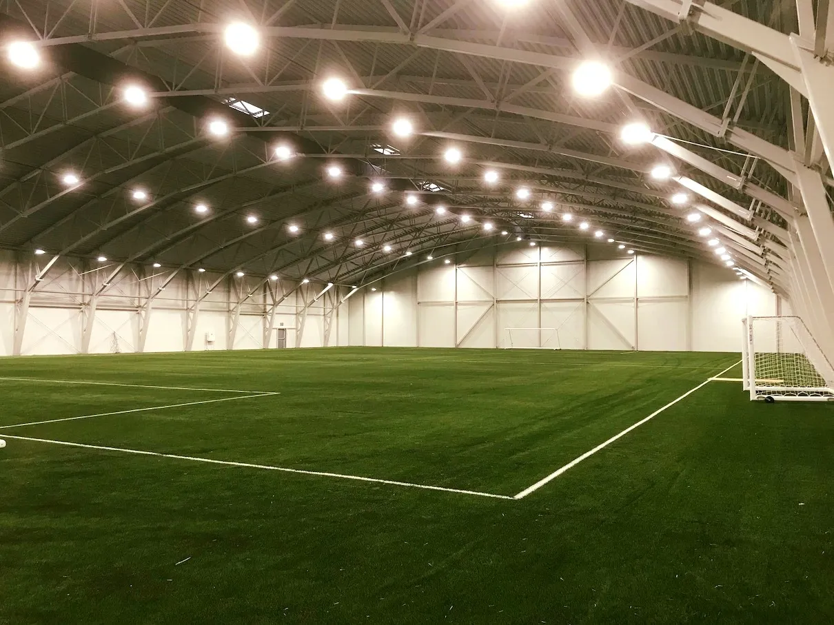 Innendørs kunstgressbane til fotball med belysning under buet tak.
