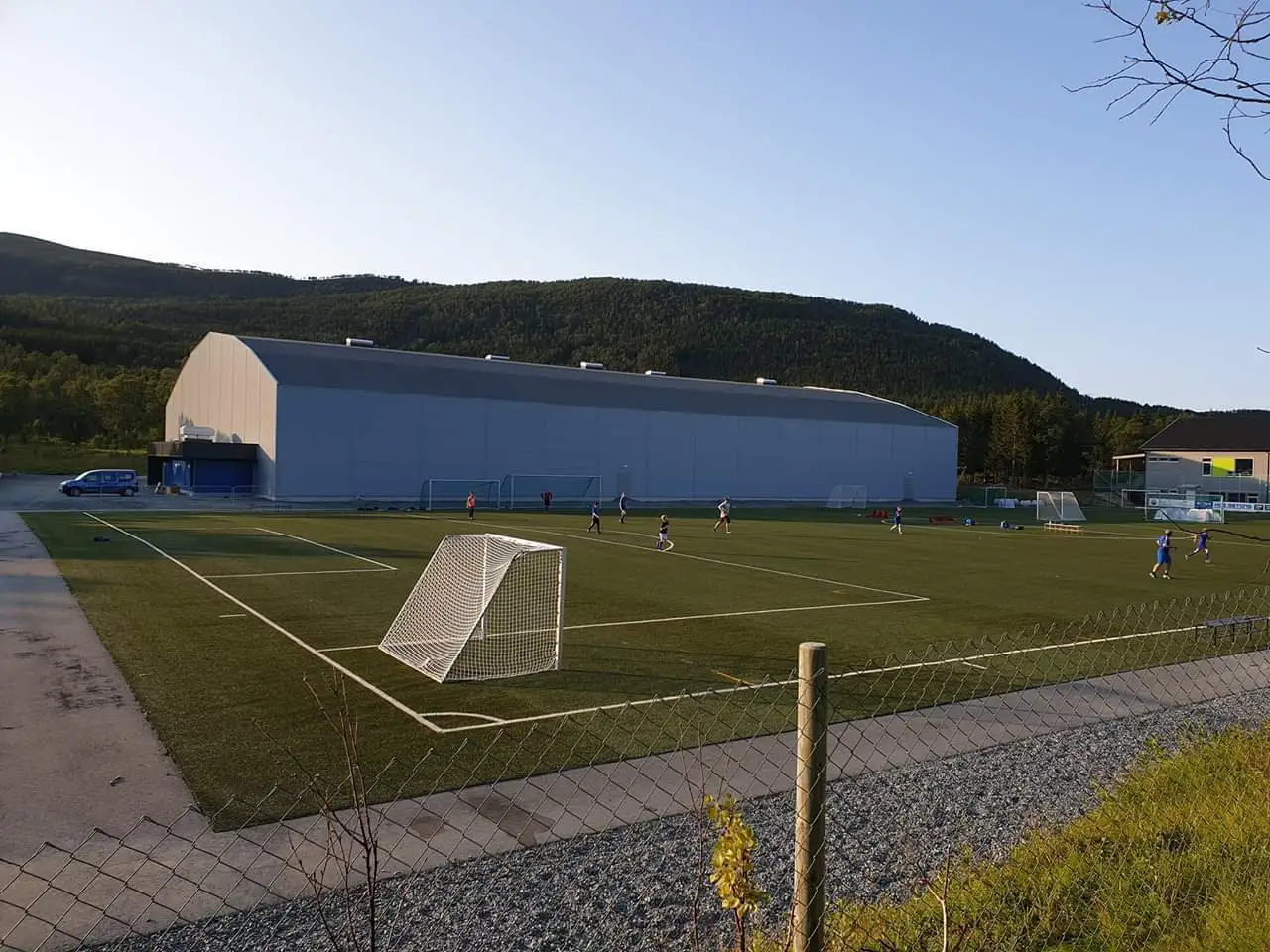 Fotballbane med barn som spiller, innhegnet med gjerde og store bygninger samt skogkledd ås i bakgrunnen.