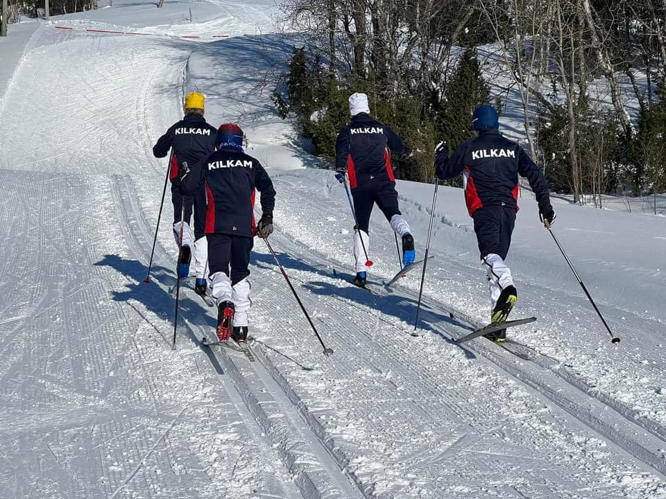 Fire personer på ski i mørke jakker med KILKAM-tekst, går opp en snødekt bakke i et skogsområde.