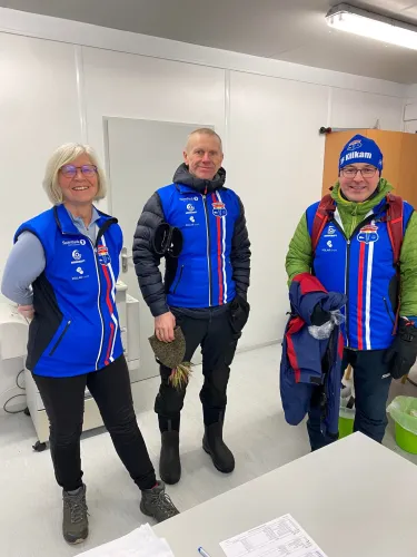 Tre personer i blå sportsjakker står innendørs foran en hvit vegg og et bord med papirer.