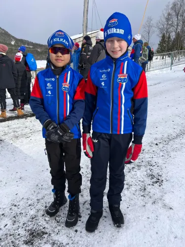 To barn kledd i blå, røde og hvite vinterjakker og luer står på snødekt bakke utendørs.
