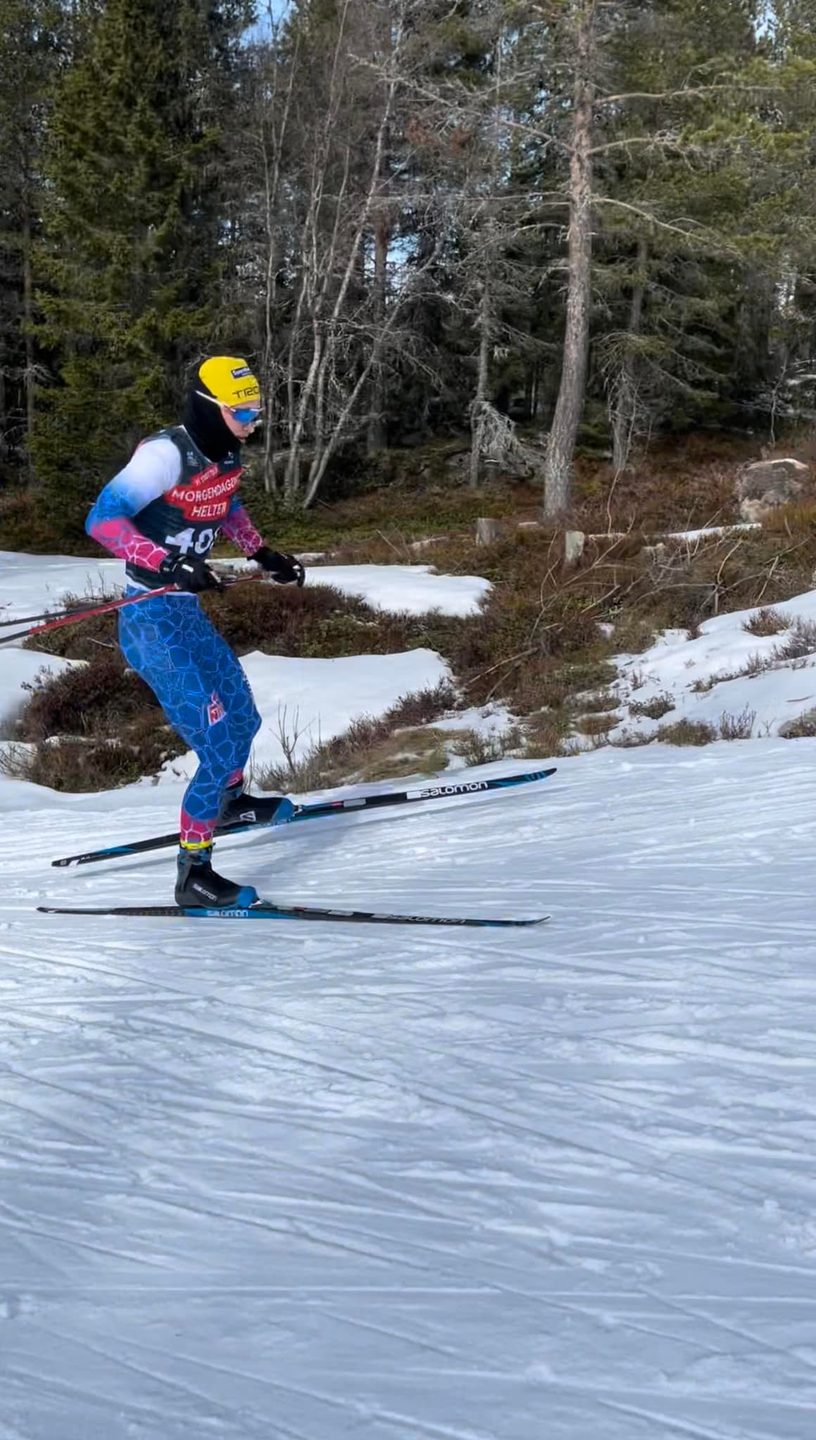 Langrennsløper i blått og rosa utstyr med gule lue på snødekt løype ved skog.