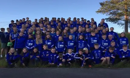 Stor gruppe barn og ungdom i blå sportsklær poserer ute på gress med trær i bakgrunnen.