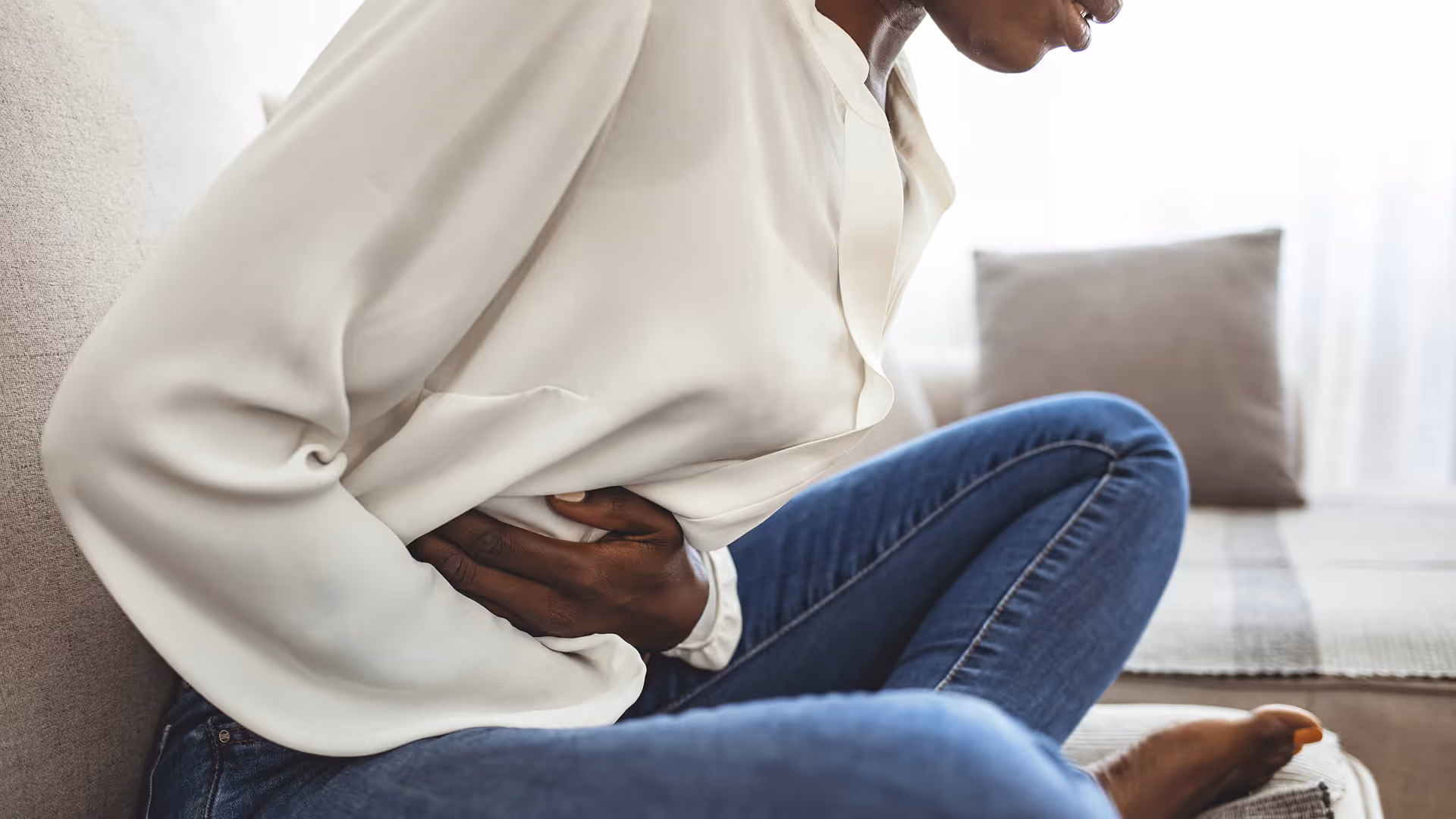 Woman sitting on a couch holding her lower abdomen in discomfort, representing fibroids and pelvic pain treated by the best gynecologic surgeon for fibroids in Los Angeles, CA.