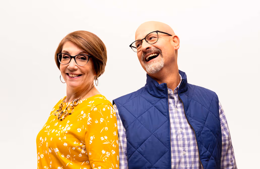 Smiling middle-aged woman in yellow floral dress and glasses standing back-to-back with a laughing bald man wearing glasses, a blue quilted vest, and a checkered shirt.