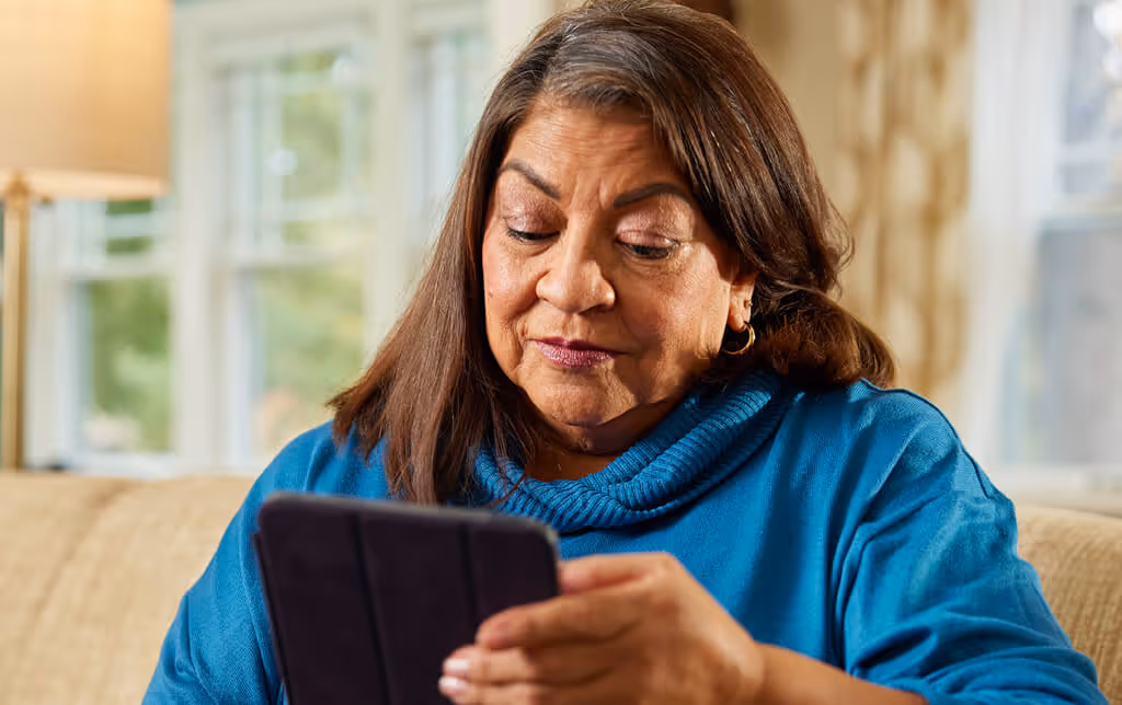 Middle-aged woman in a blue sweater sitting on a couch looking down at a tablet indoors.