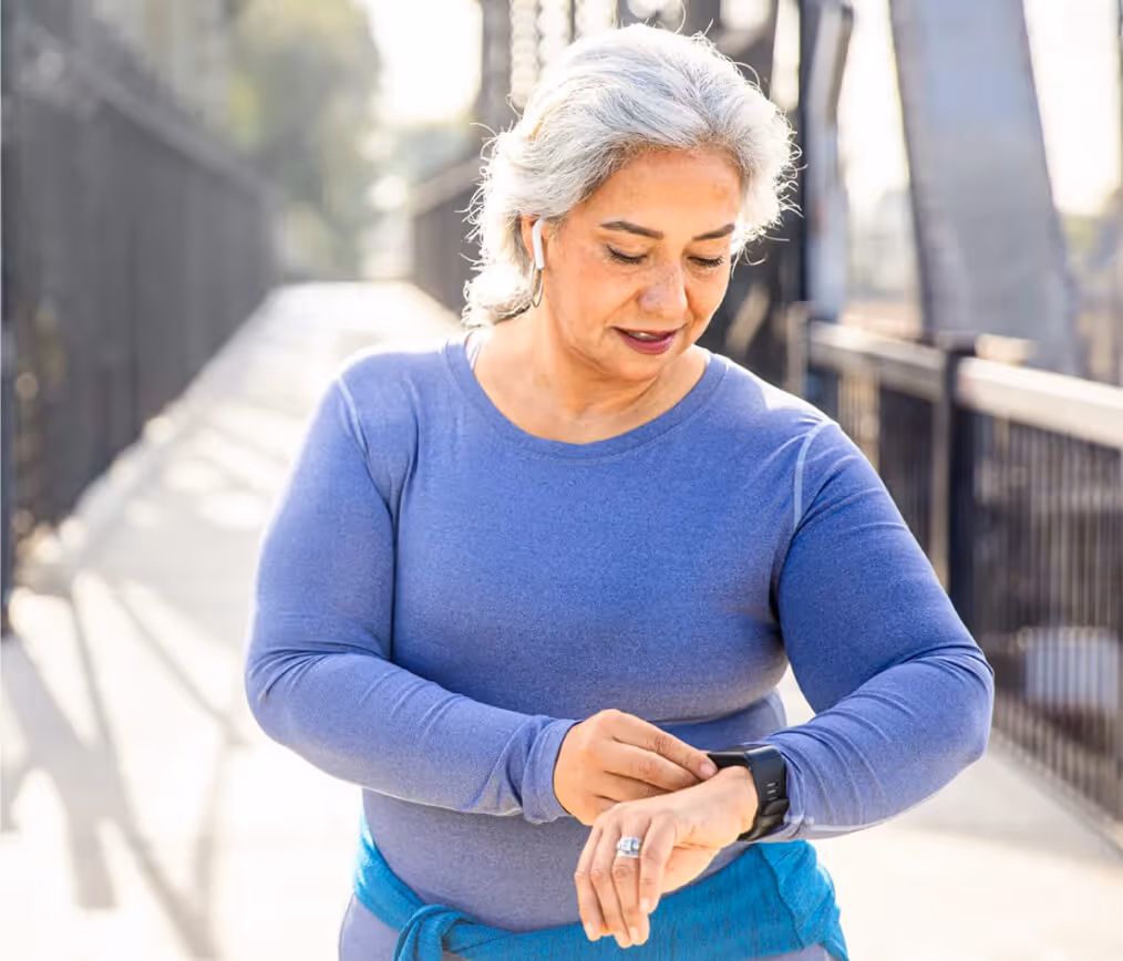 Mature woman in blue activewear looking at her smartwatch while walking outdoors on a sunny day.