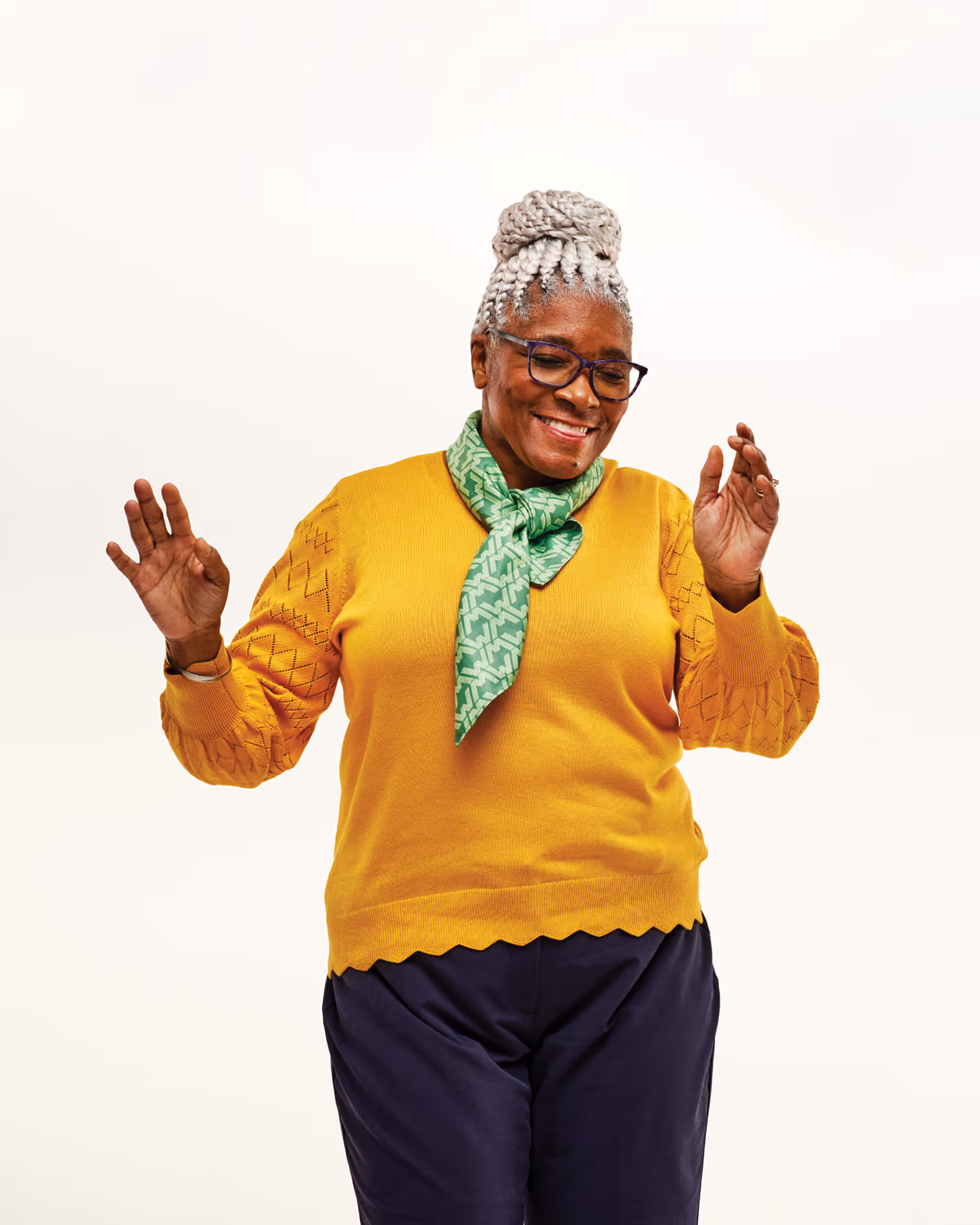 Smiling older woman with braided gray hair wearing glasses, a yellow sweater, green scarf, and dark pants, dancing with hands raised.