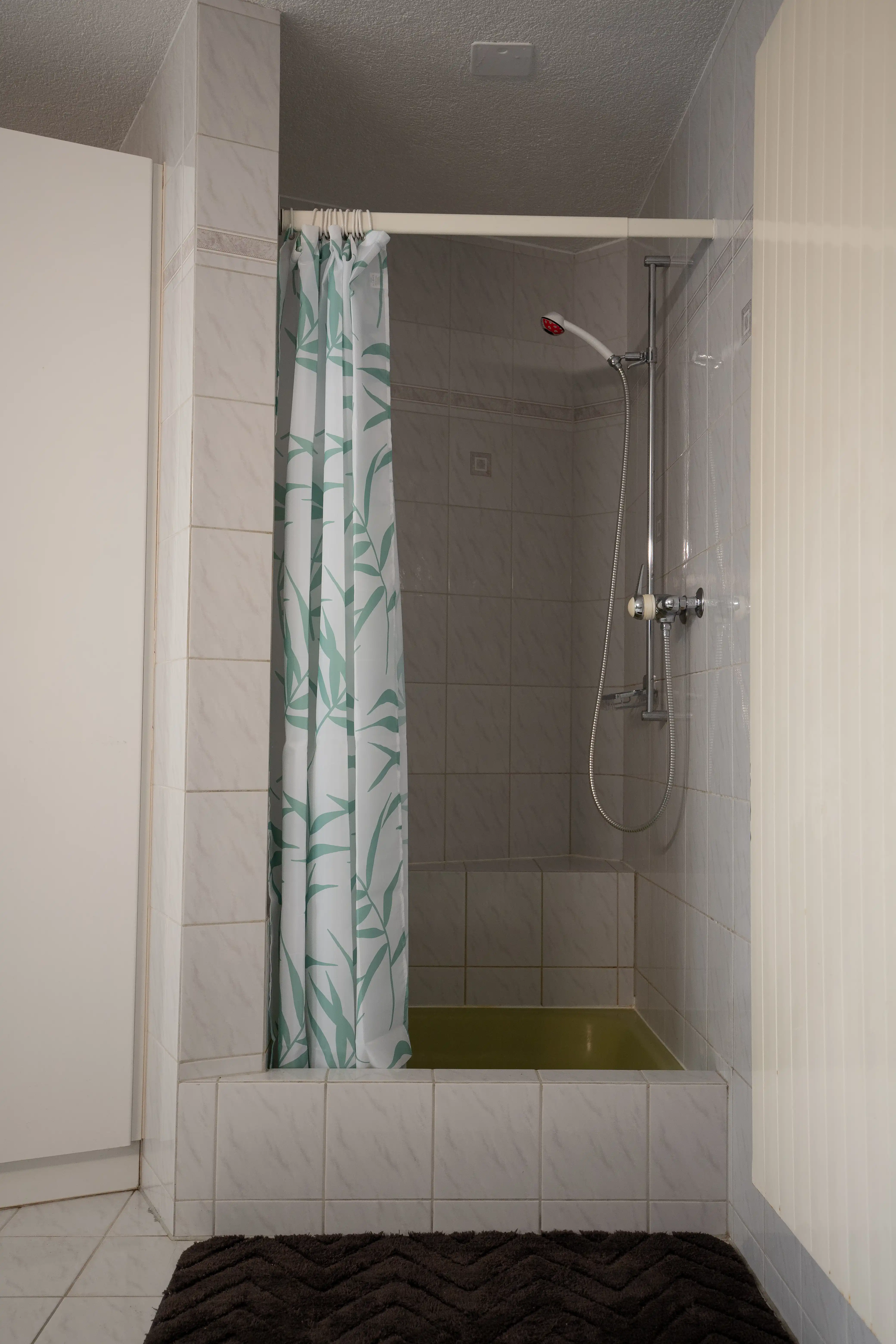 Geflieste Dusche mit Vorhang, Handbrause und erhöhtem Einstieg in hellem Badezimmer